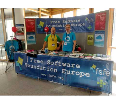 Two volunteers behind a booth of the FSFE presenting information and merchandise material.